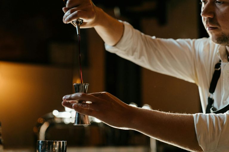 A bartender in a uniform expertly measures spirits with a jigger in a dimly lit indoor bar setting.