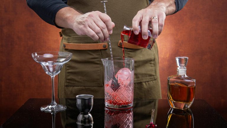 A bartender mixes a classic cocktail with red liquor, ice, and stirring implements.