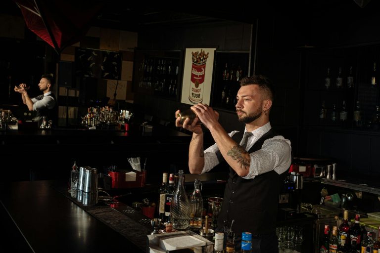 Skilled bartender shaking a cocktail mixer at a bar counter, surrounded by spirits and bar tools.